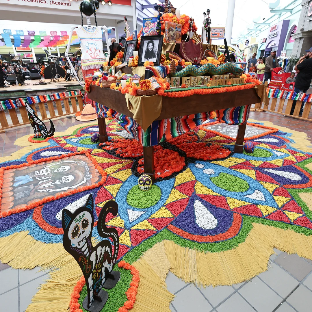 Organizer-selected reference photo of a Día de Muertos altar and colorful floor decorations inside La Gran Plaza; photograph date not specified.